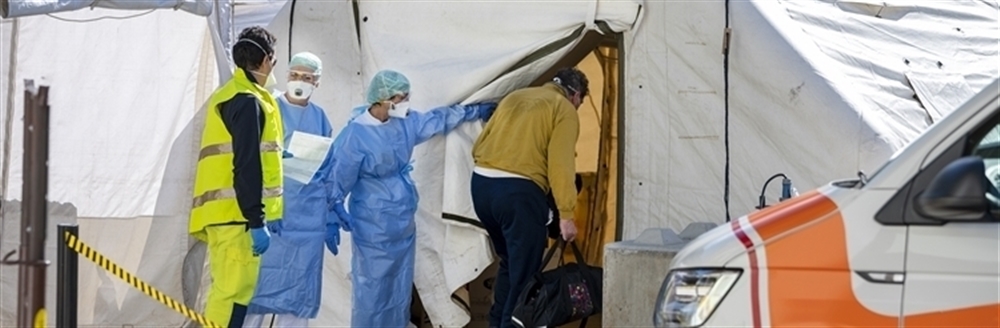 A medical professional in scrubs and full PPE holds open a triage tent entrance for someone entering. Another medical professional and a first responder in reflective attire stand nearby, talking. The front of an ambulance is in the foreground.